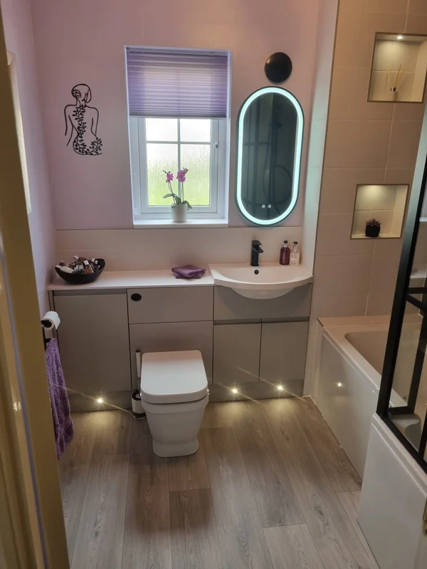 Blush pink bathroom with a fitted vanity unit, illuminated oval mirror, wood-effect flooring and low-level floor lighting
